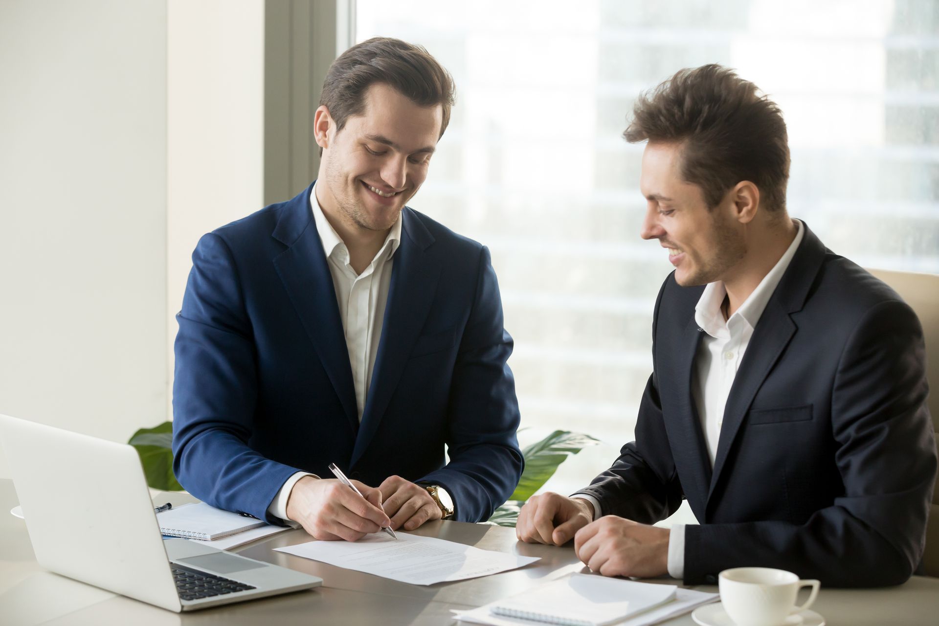 Two men in suits smiling, one signing a document at a desk with a laptop and coffee cup.