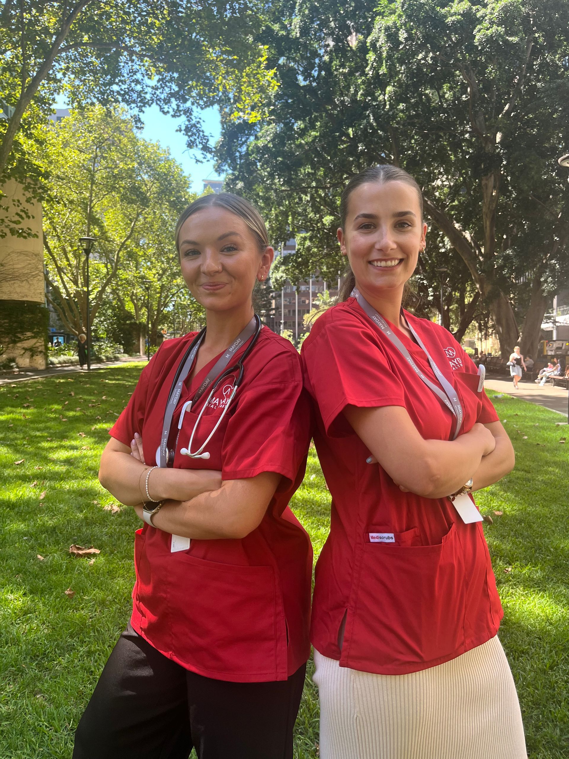 Two women in red scrubs stand back-to-back with arms crossed in a park setting.