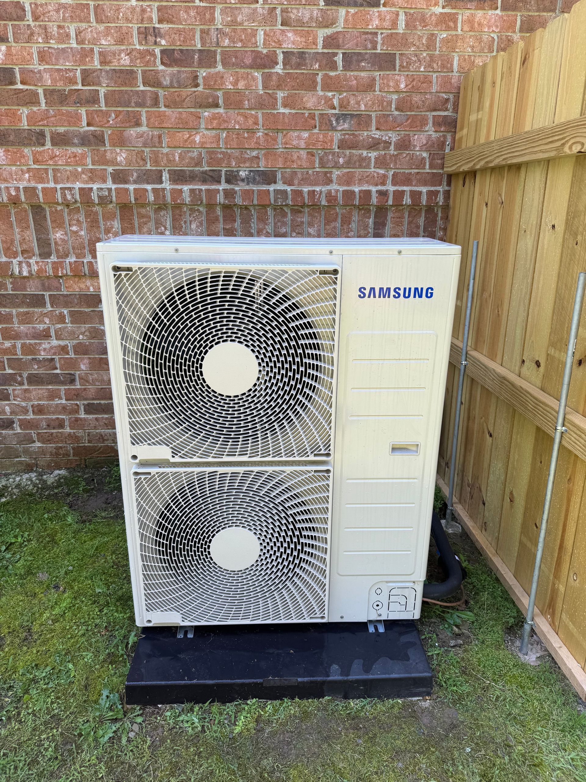 A samsung air conditioner is sitting in front of a brick wall.