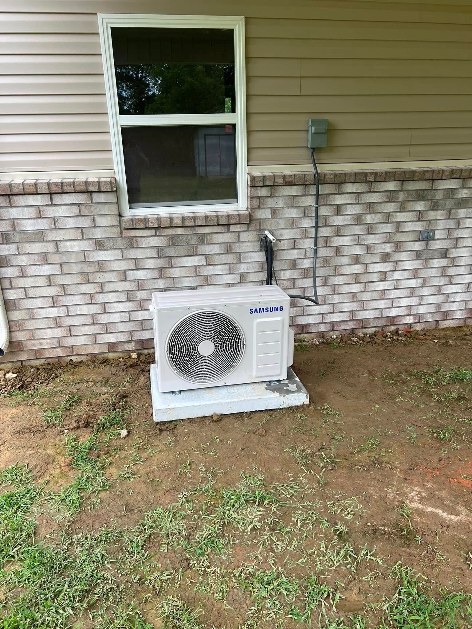 A white air conditioner is sitting in the grass in front of a brick wall.