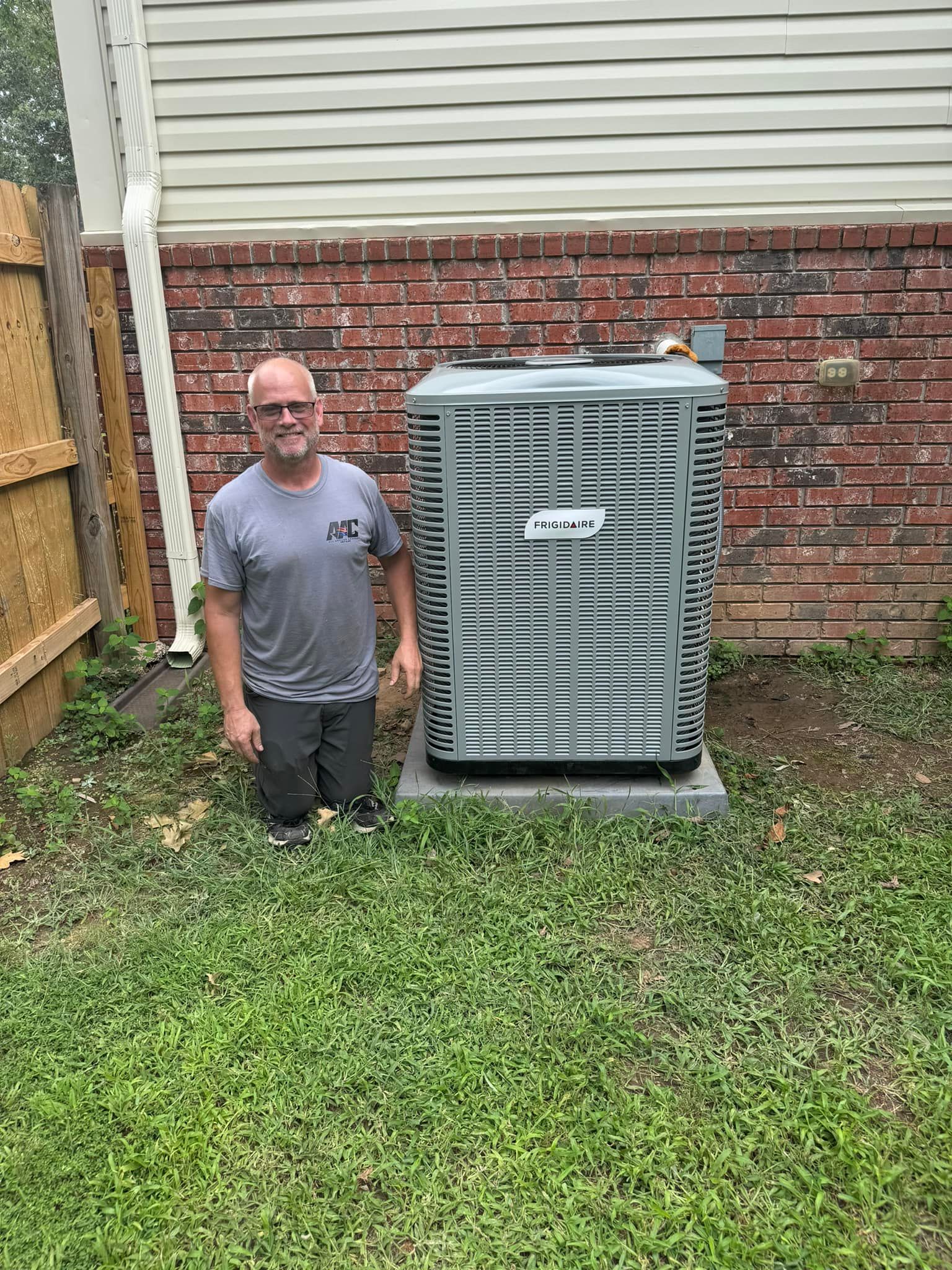 A man is kneeling in front of an air conditioner in a backyard.