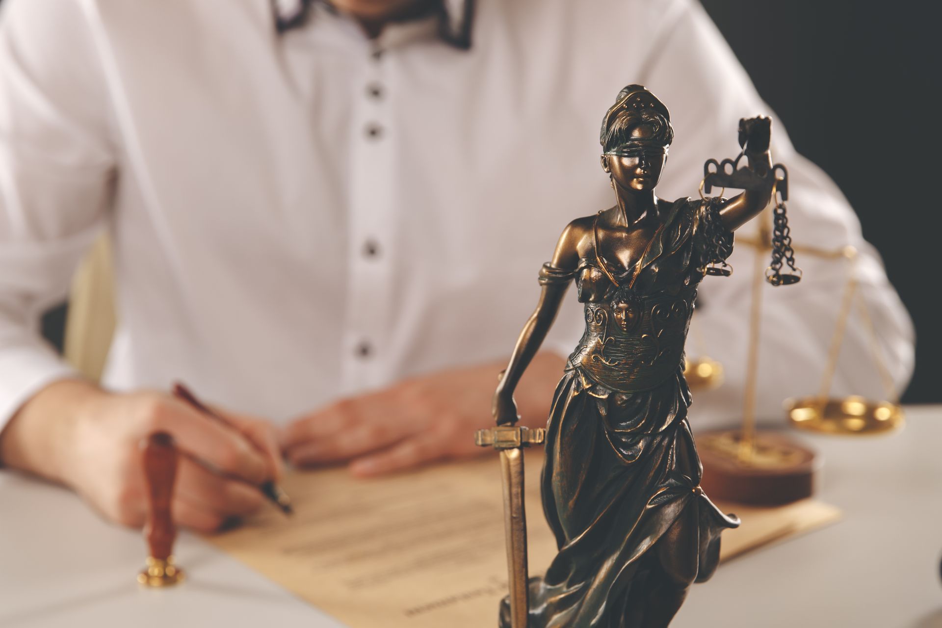 A person signing a document at a desk with a Lady Justice statue in the foreground.