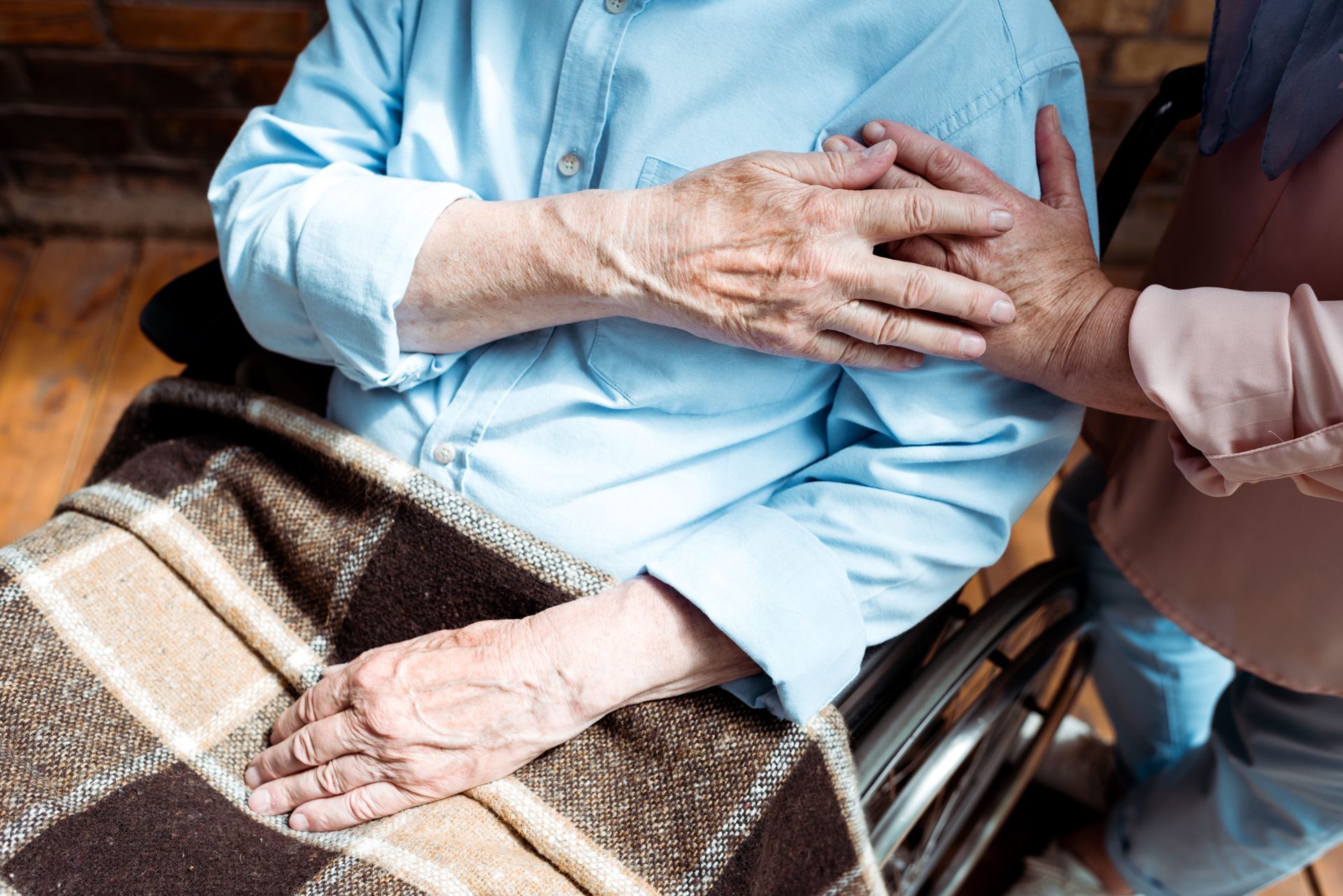 Two people hold hands over a brown blanket in a wheelchair, suggesting comfort and support