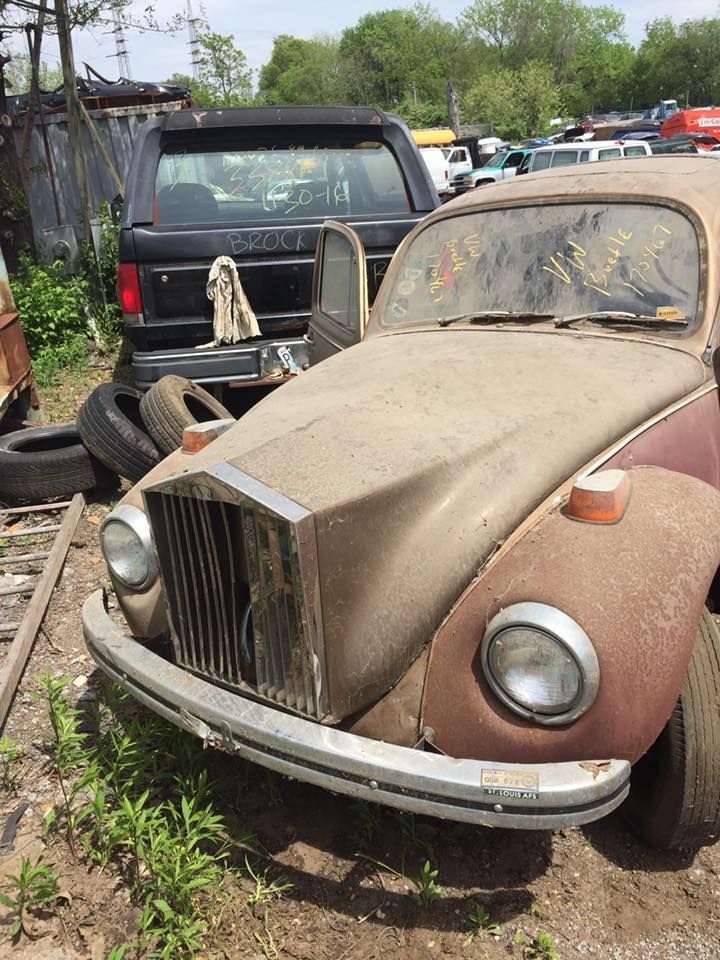An old rusty car is parked in a junkyard.