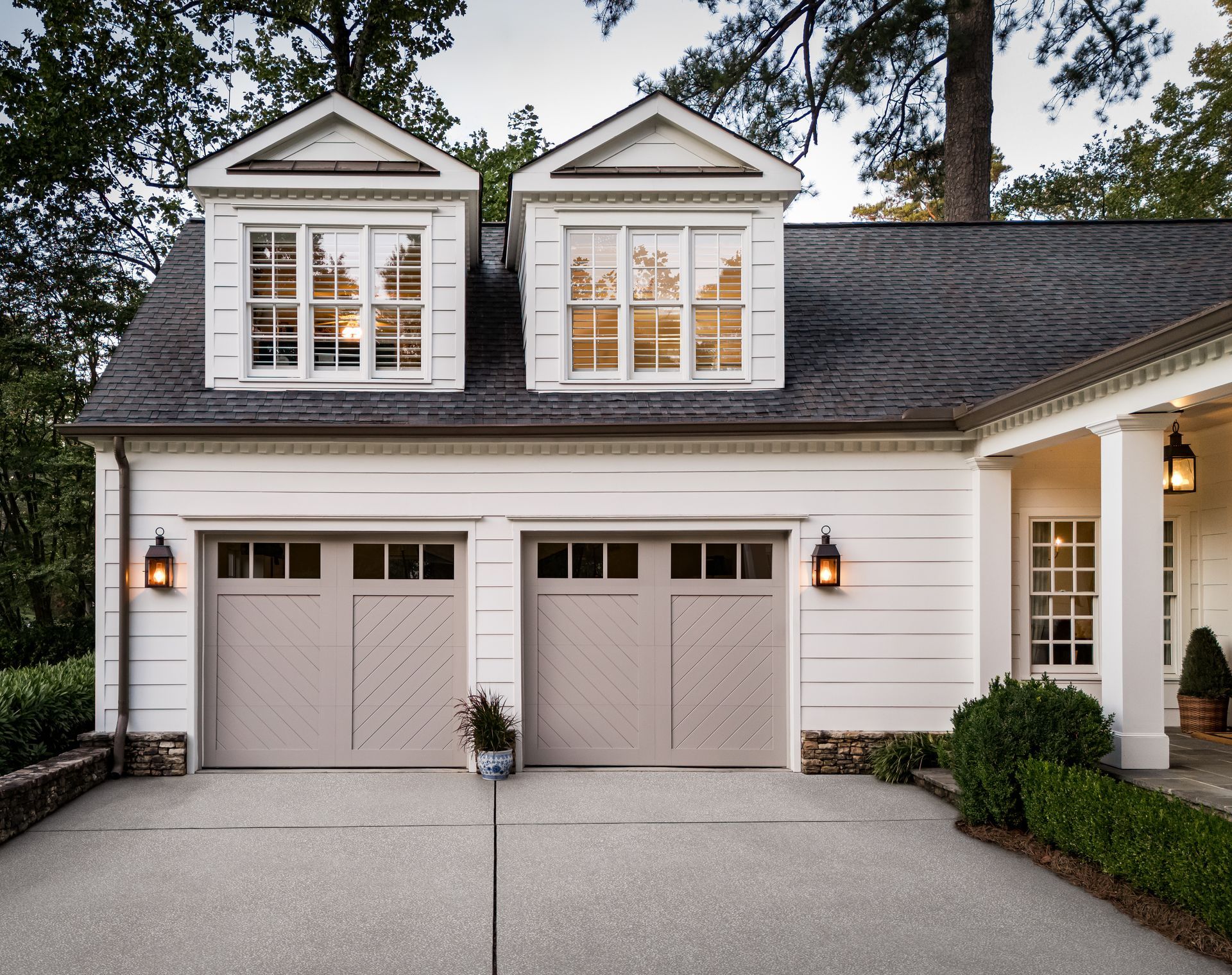 Tan garage doors with two windows above, set in a white house, gravel driveway.