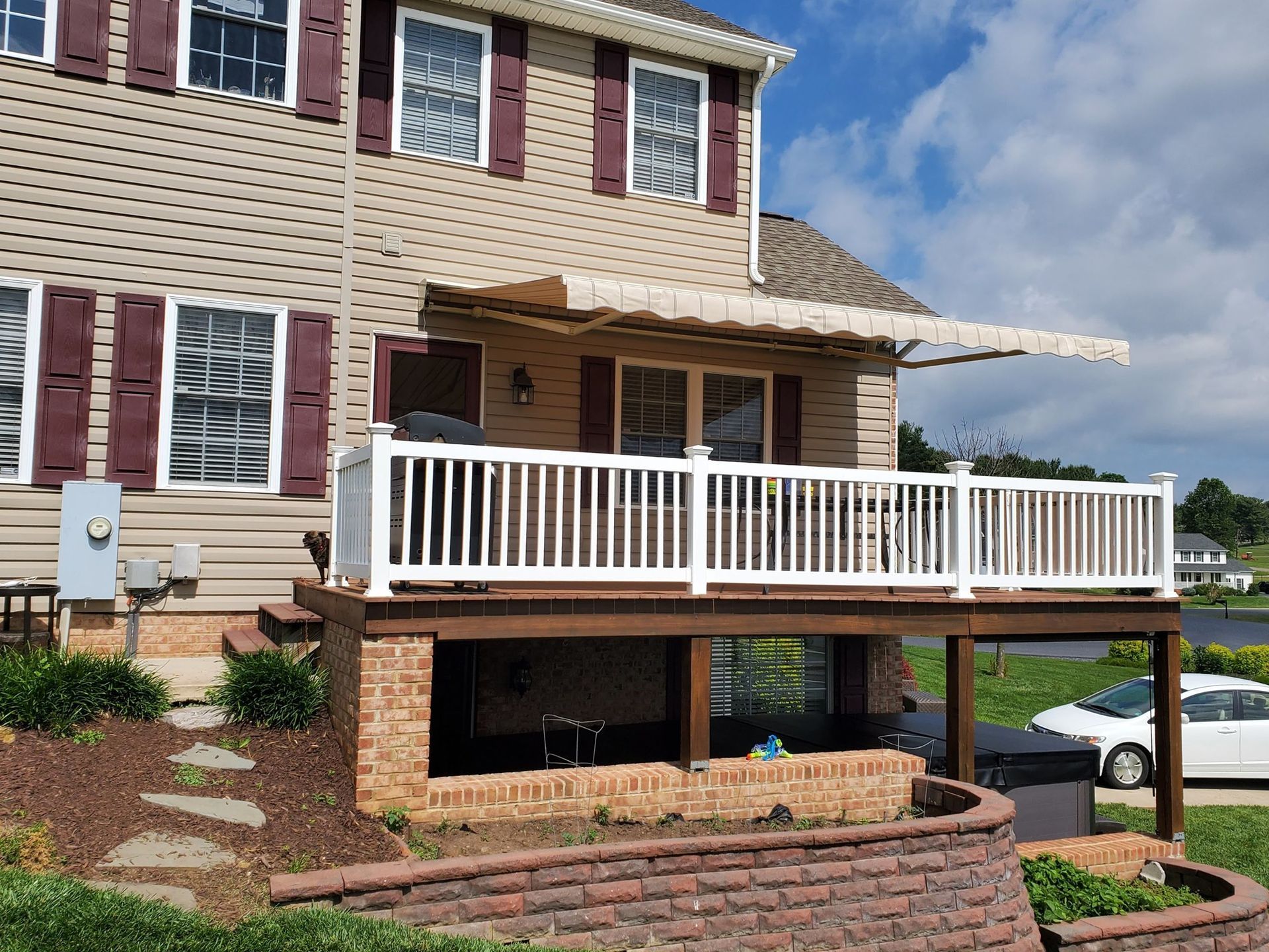 Two-story house with a white deck and awning. Red brick retaining wall in front, car to the right.