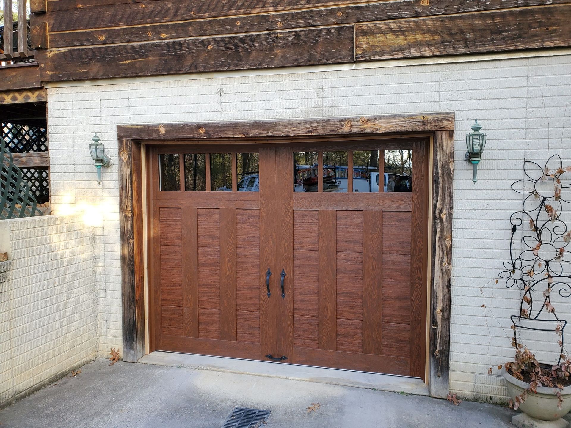 Brown garage door with windows, rustic wooden trim, on white brick building. Two sconces and a plant on either side.