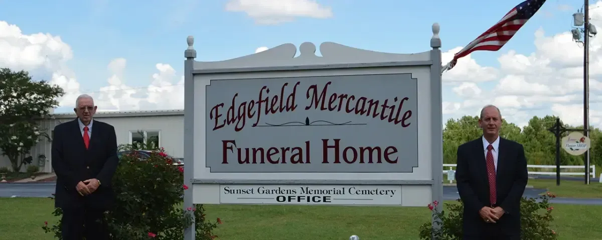 Two men in suits stand near a sign for Edgefield Mercantile Funeral Home. An American flag waves above.