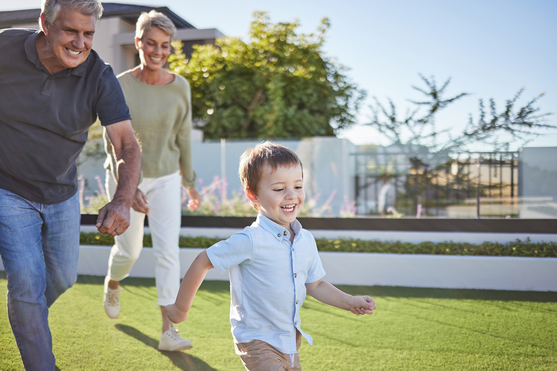 A child runs on grass, smiling, holding hands with an older man as an older woman walks behind them. Sunny outdoors.