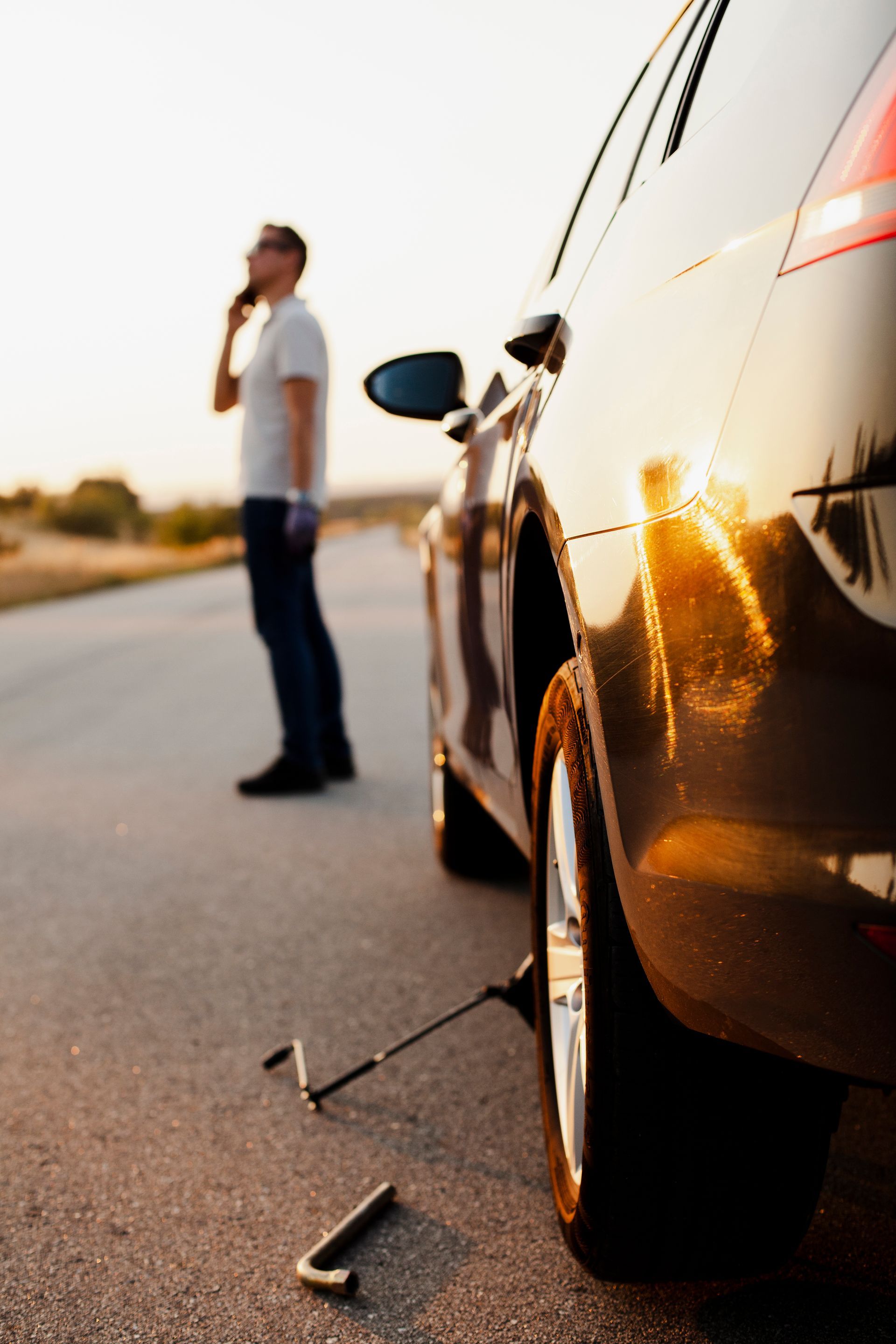 Man on phone by car with jack on a roadside at sunset.
