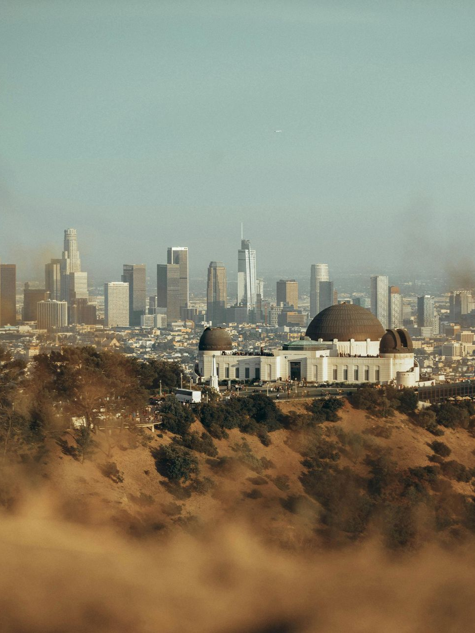 Griffith Observatory overlooks Los Angeles skyline, framed by brownish hills and hazy sky.