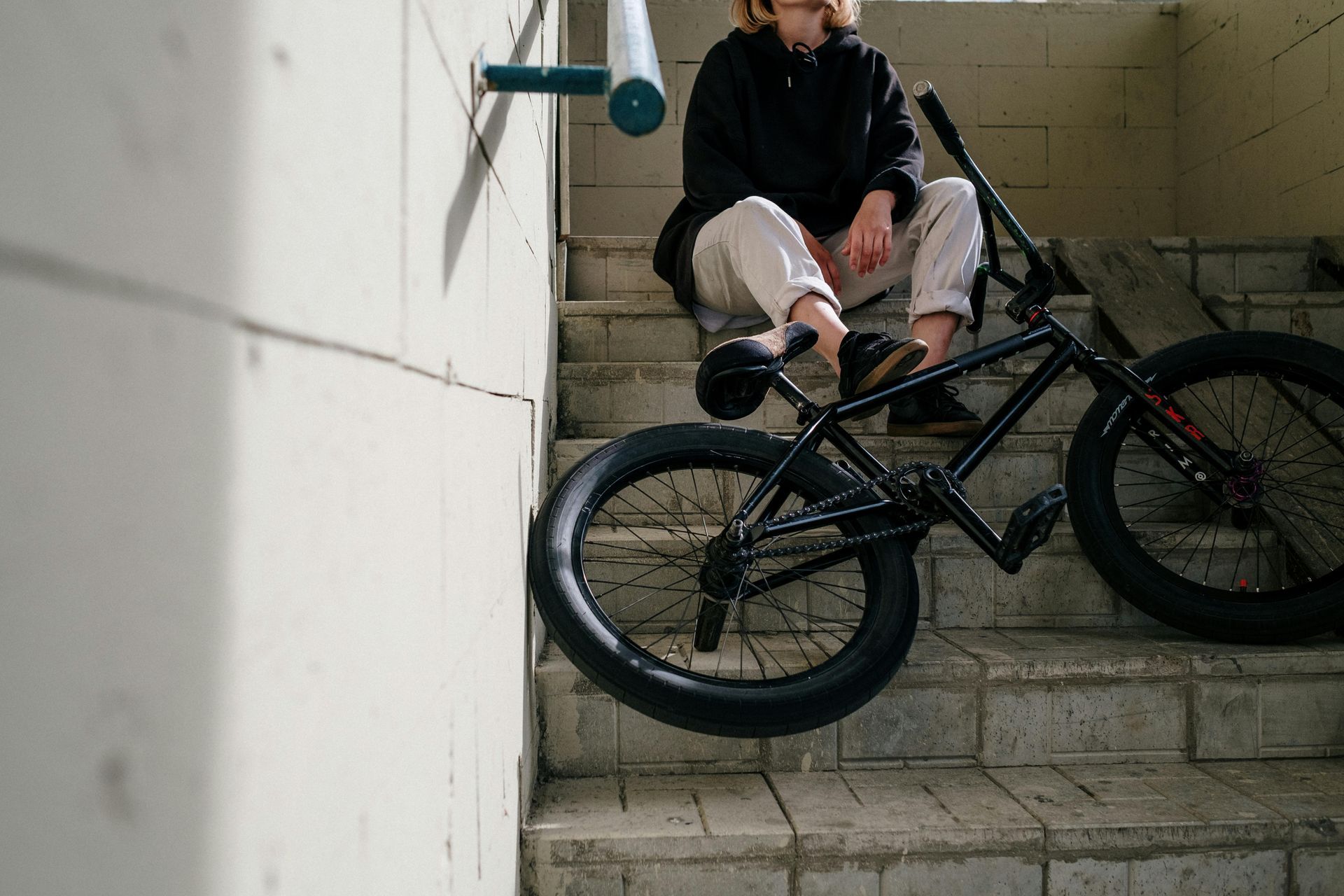 Person in black hoodie and white pants sits on concrete stairs with a black BMX bike.