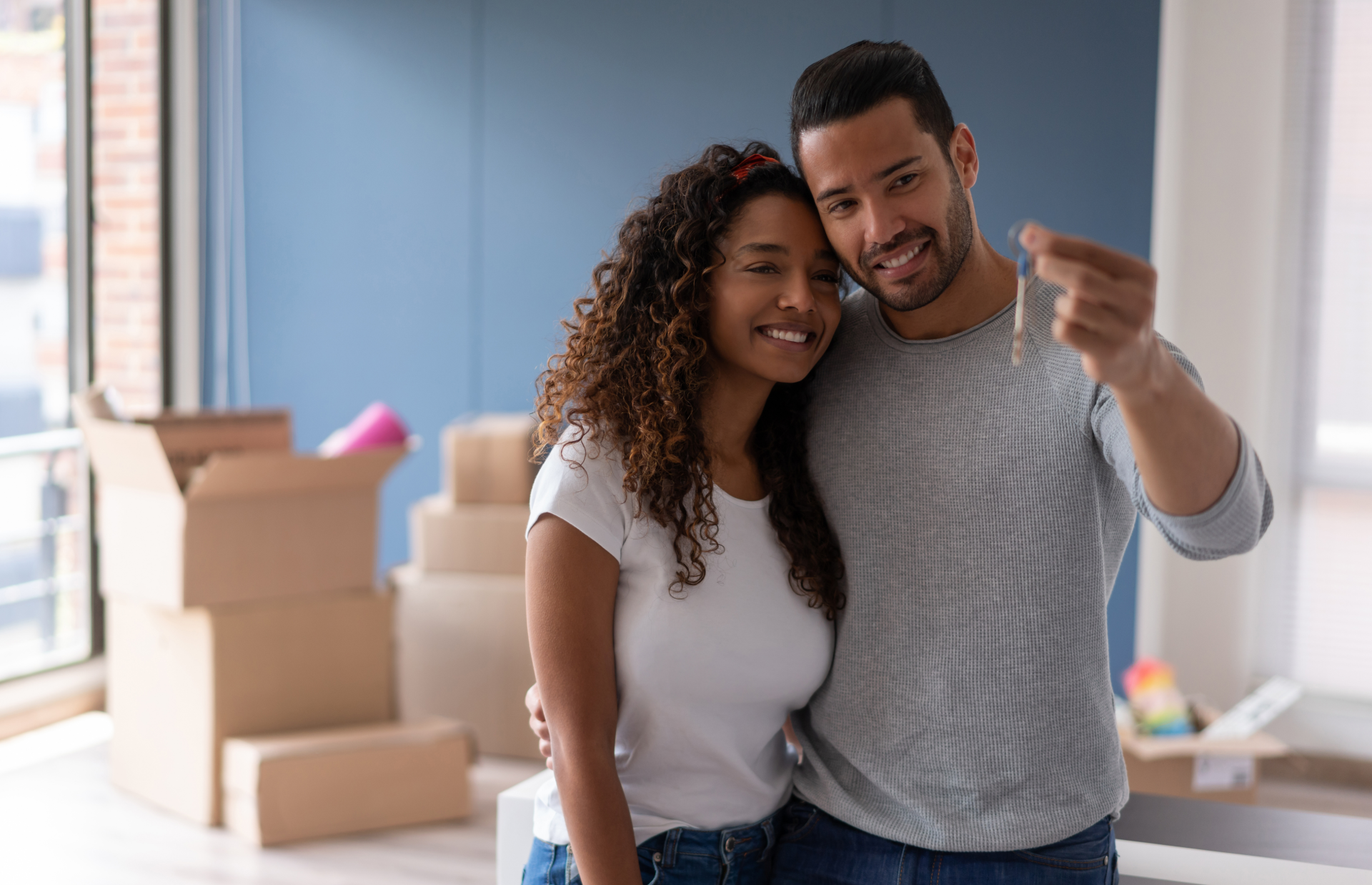 A man and a woman are posing for a picture in their new home . the man is pointing at the keys.