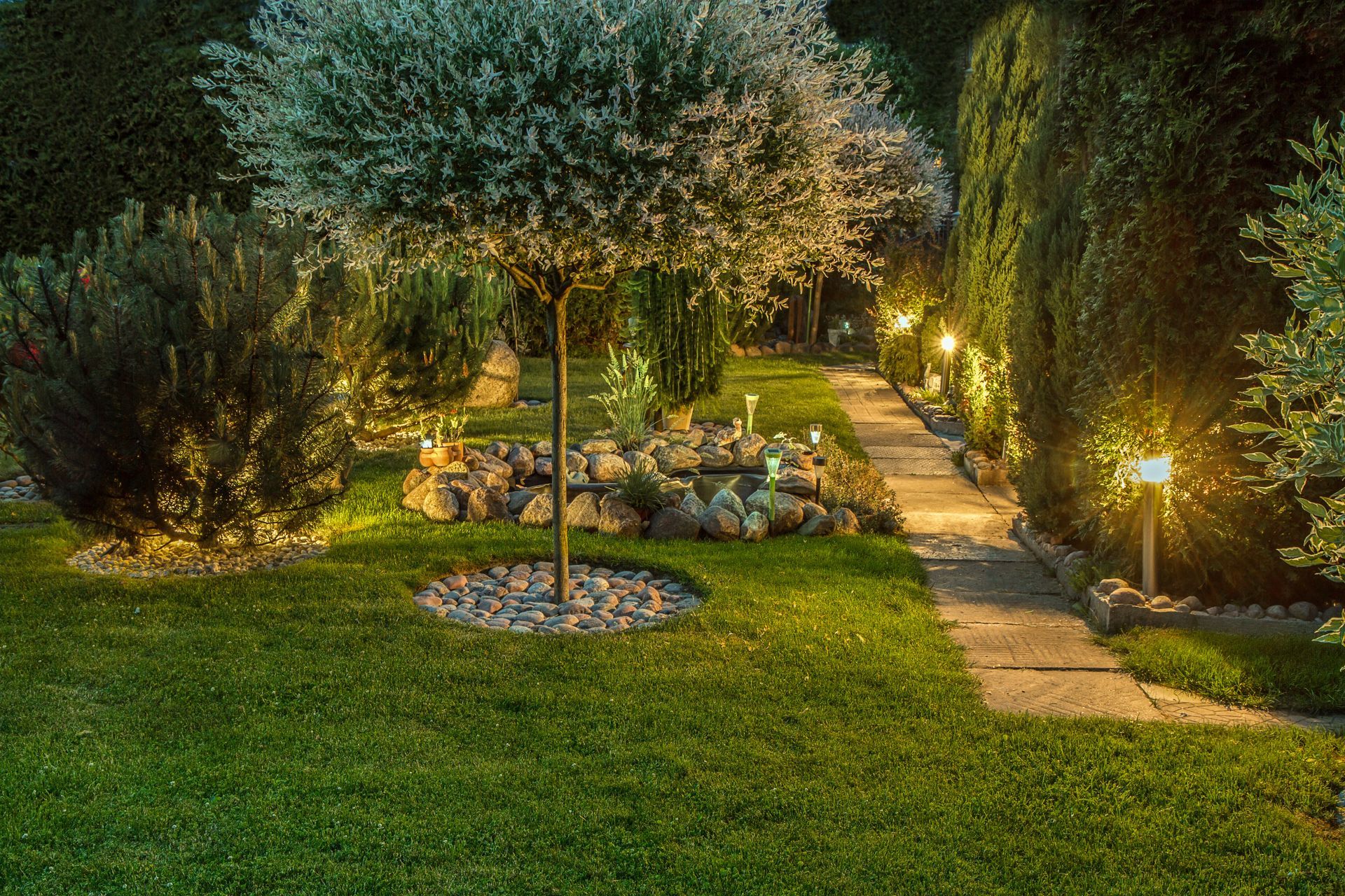Nighttime garden with lit pathways, trimmed lawn, and trees surrounding a small seating area