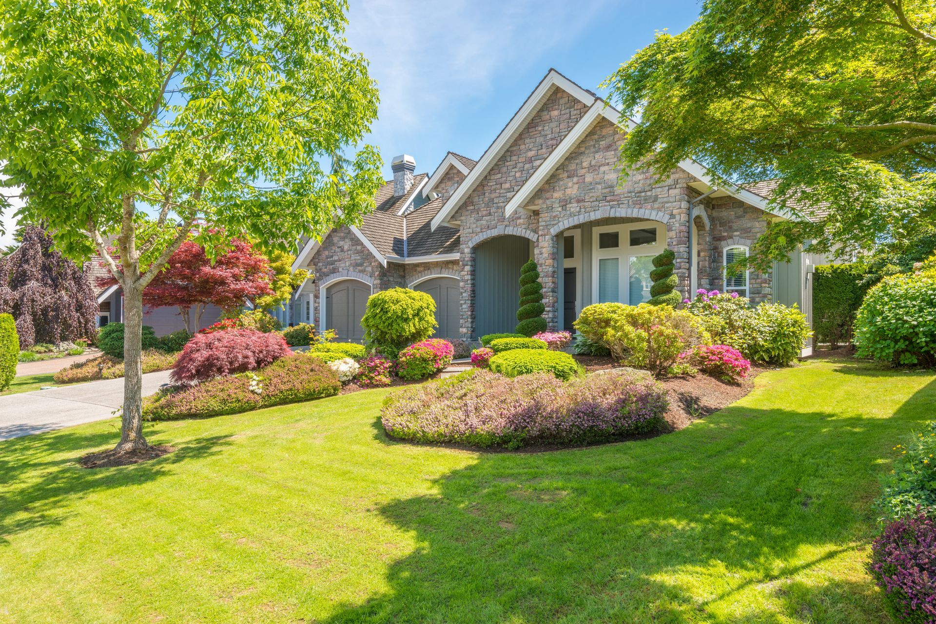 Sunny suburban brick house with manicured lawn, flowering shrubs, and green trees