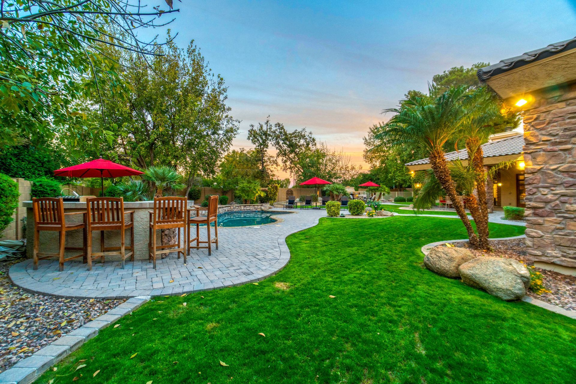 Backyard patio with wooden dining set, curved pool, lush lawn, and tropical landscaping at sunset