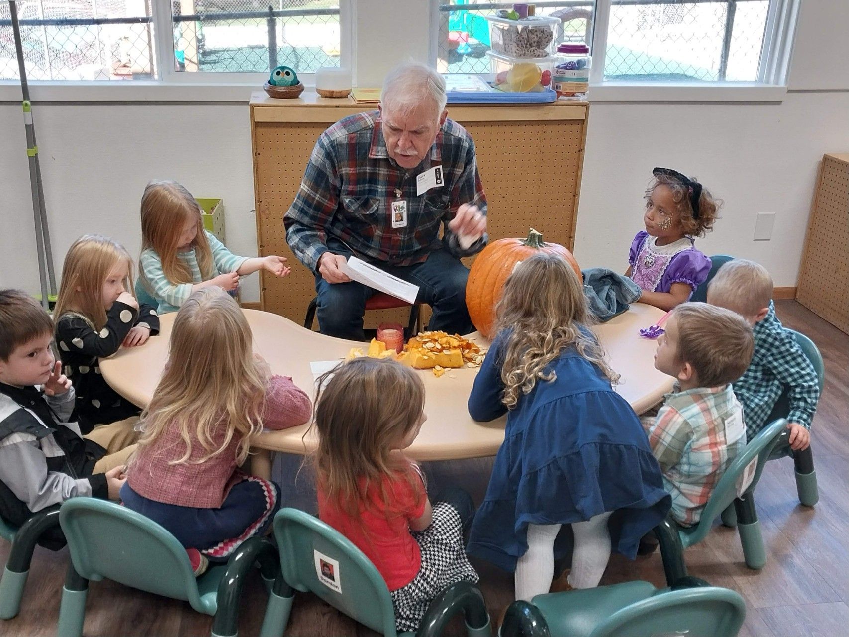 An adult sits with children at a table with a pumpkin and snacks, sharing paper during a classroom activity.