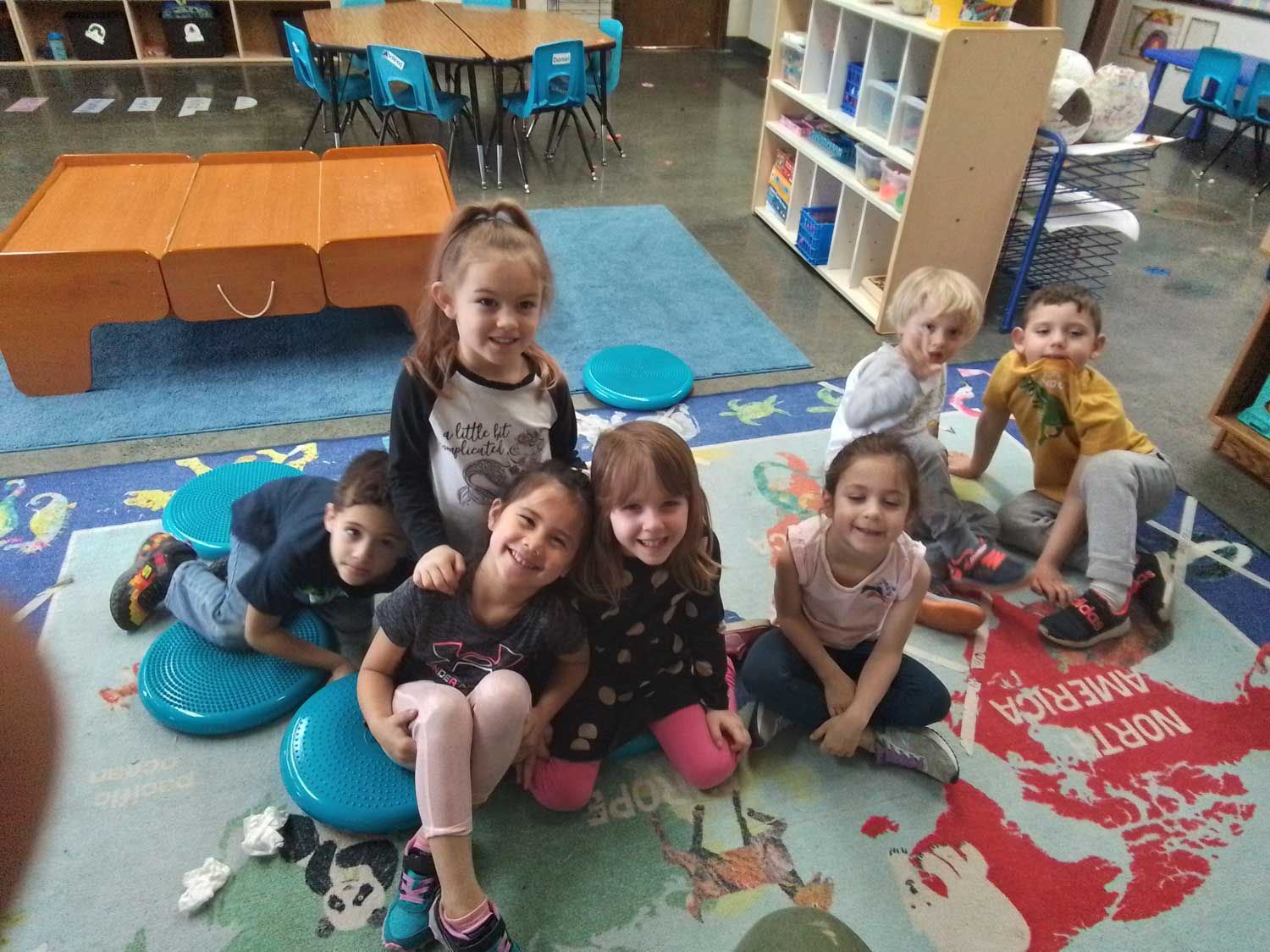 A group of eight children sitting on a colorful floor map in a classroom, smiling and posing for a photo.
