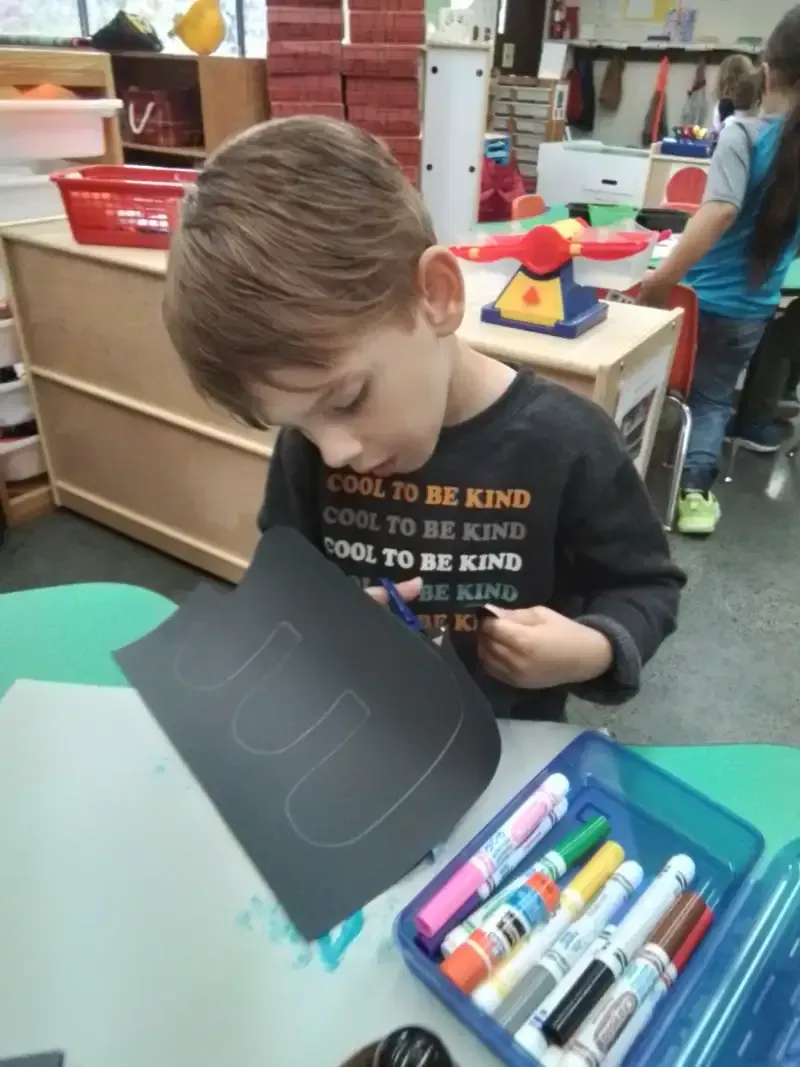 A child uses scissors to cut a letter shape out of black construction paper at a classroom table with markers nearby.