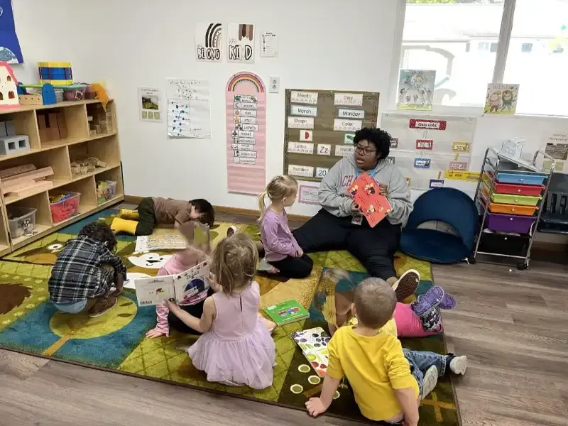 An adult sits on a rug with five young children in a classroom, reading from a colorful book.