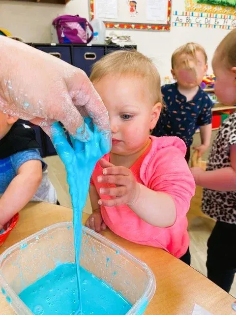 A person holds blue slime over a container while a child reaches out to touch it in a classroom.