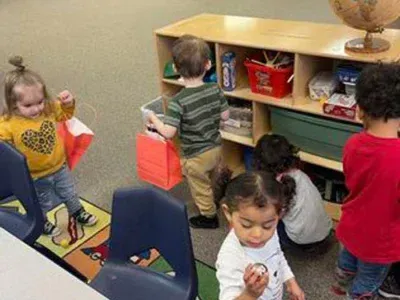 A group of children in a classroom carrying bags and engaging with classroom materials near a toy shelf.