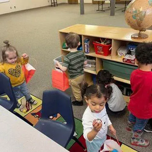 Children in a classroom setting playing with toys, including bags and a globe on a shelf.