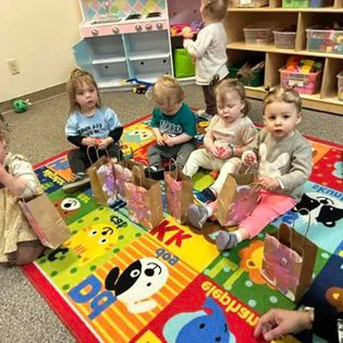 Six children sit on a colorful alphabet rug in a playroom, each with a small brown gift bag in front of them.