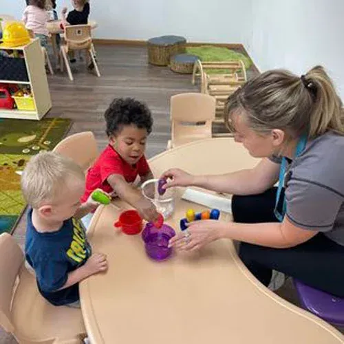An educator sits at a table with two children, playing a color-matching game with plastic cups and small items.