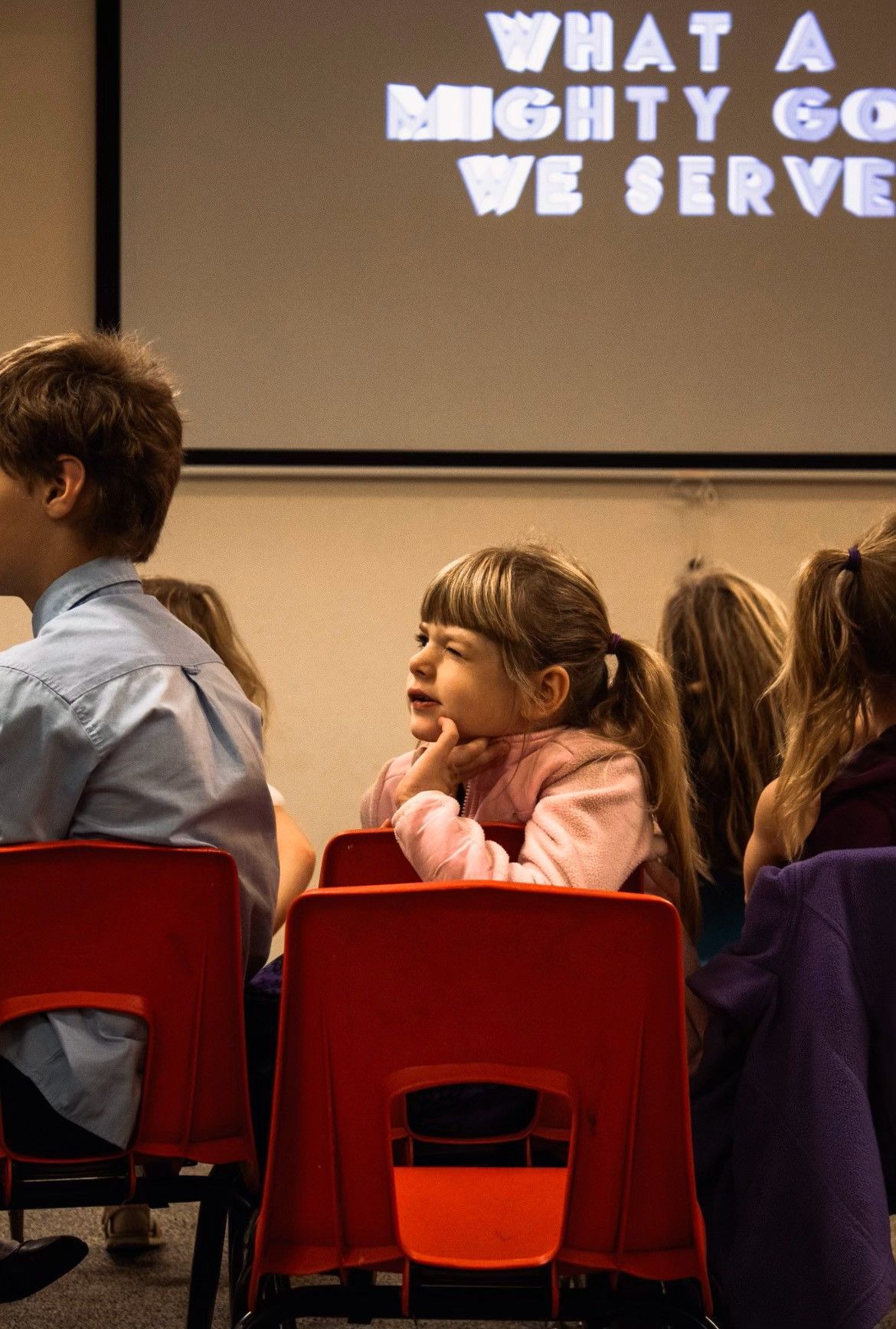 Children sit on red chairs in a room with a projection screen displaying 
