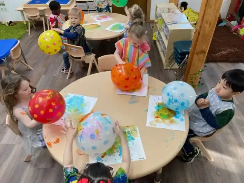 A group of children sits at tables in a classroom, using balloons to paint on paper with colorful polka-dot patterns.