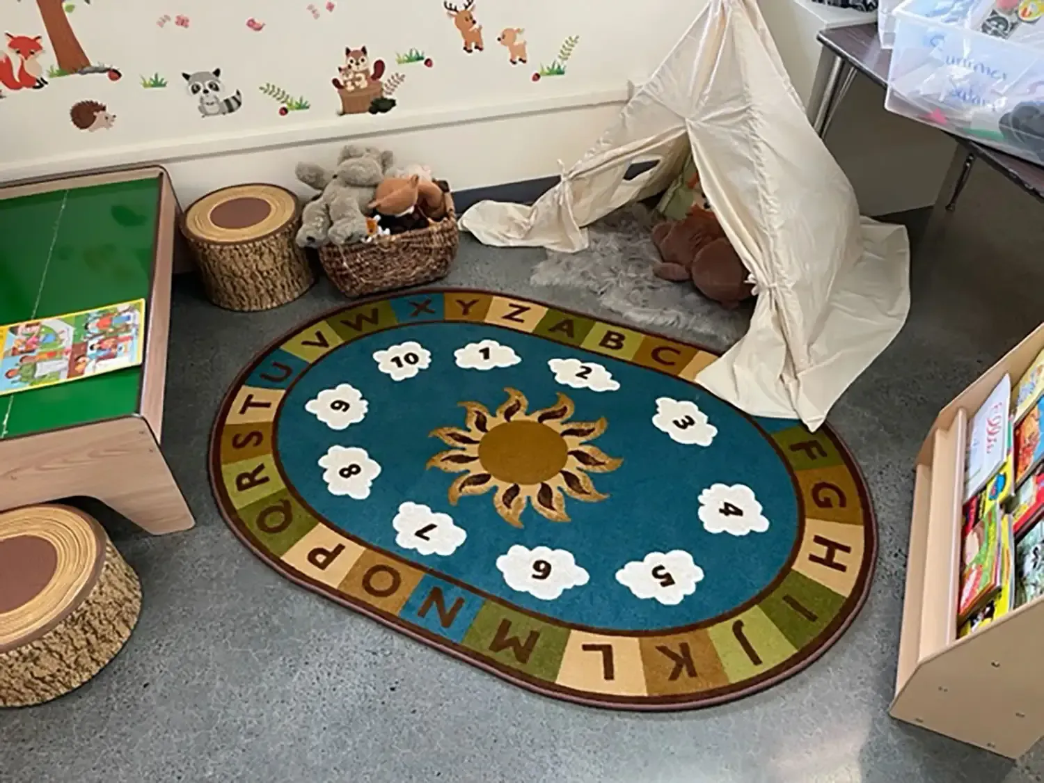 A play corner with a small teepee, an alphabet and number rug with a sun center, toy storage, and log-shaped seats.