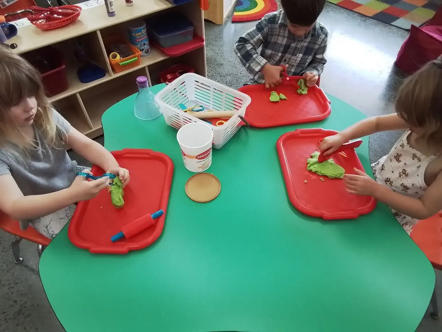 Three children sit at a green table, using red trays and tools to play with green modeling dough in a classroom.