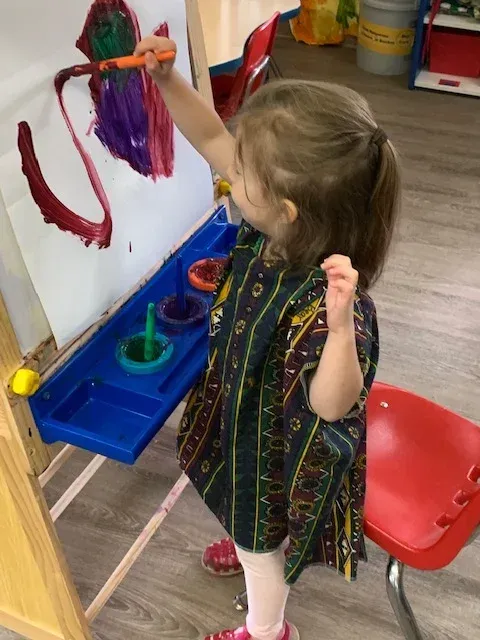 A child wearing a patterned smock paints red and purple swirls on paper attached to an easel in a classroom.