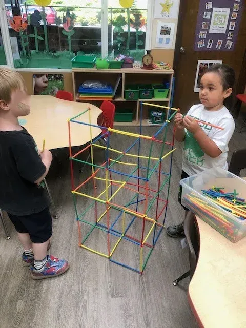 Two children build a tall, colorful geometric structure out of plastic sticks and connectors in a classroom setting.