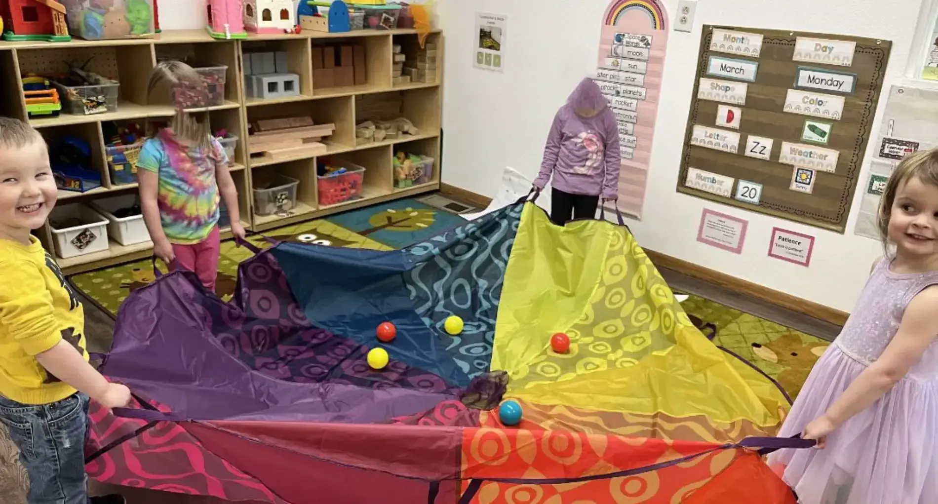 Children in a classroom hold a multi-colored parachute with balls on top while smiling and participating in play.