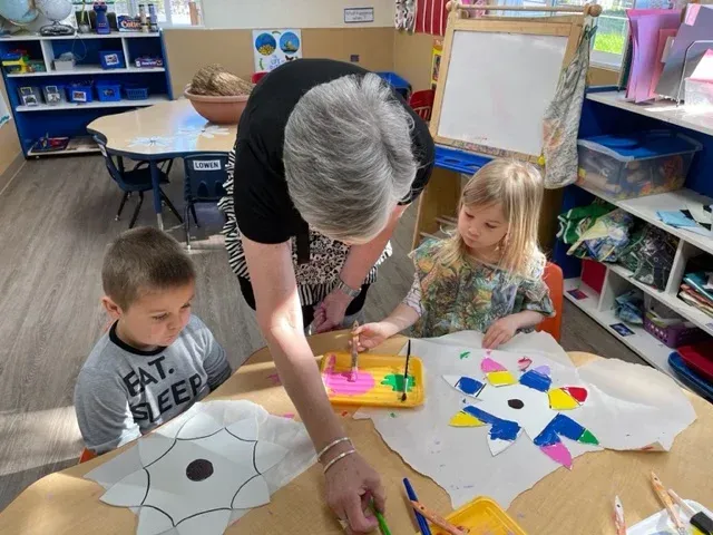 A teacher and two children paint star-shaped paper cutouts at a table in a classroom.