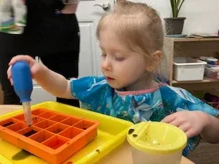 A child uses a blue dropper to transfer liquid from a yellow cup into an orange grid tray on a yellow surface.