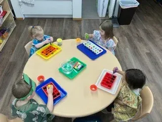 Four children sit around a round table in a classroom, using droppers to transfer colored water into ice cube trays.