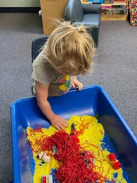 A child leans over a large blue bin filled with yellow filler and red spaghetti-like sensory materials and small toys.