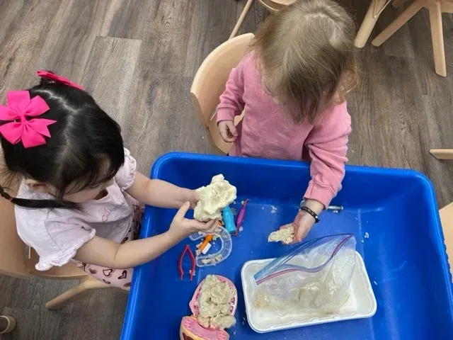 Two children play with modeling clay and plastic tools inside a blue sensory bin at a table.
