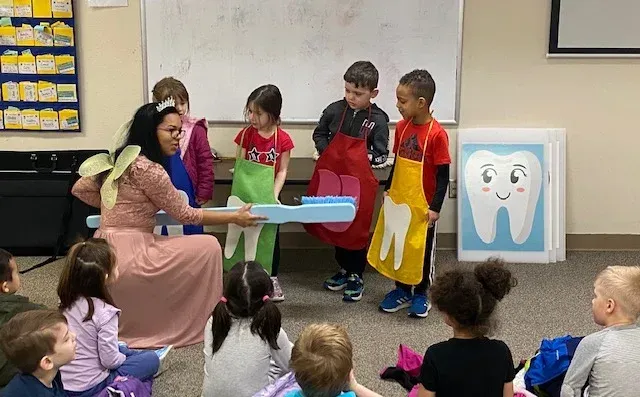 A person in a costume holds a large prop toothbrush for children wearing colorful aprons with tooth designs in a classroom.