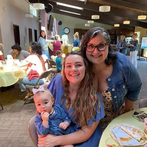 A woman holding a baby in her lap, next to another smiling woman, at a social gathering in a room with light fixtures.