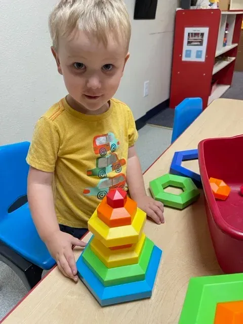 A child sits at a table stacking colorful, multi-shaped blocks into a pyramid in a classroom setting.