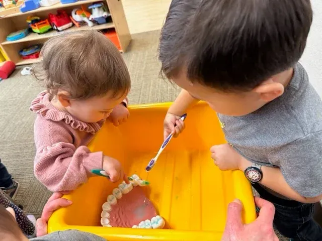 Two children play with toothbrushes and a large dental model in a yellow bin in a classroom setting.