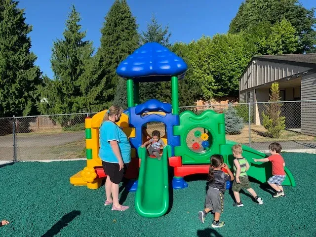 An adult stands near a small, colorful plastic play structure on a green playground with four children playing nearby.