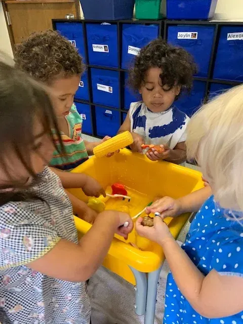 Four children play together around a yellow sensory table in a classroom with blue storage bins in the background.