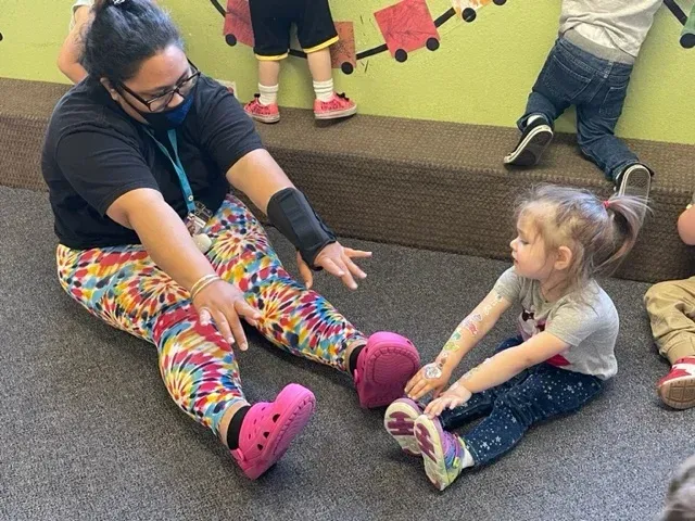 A person in tie-dye pants and a child sit on a carpeted floor, reaching toward each other in a stretching activity.