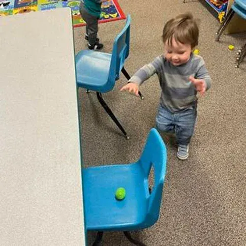 A toddler in a striped shirt reaches toward a green ball sitting on a blue plastic chair in a classroom.