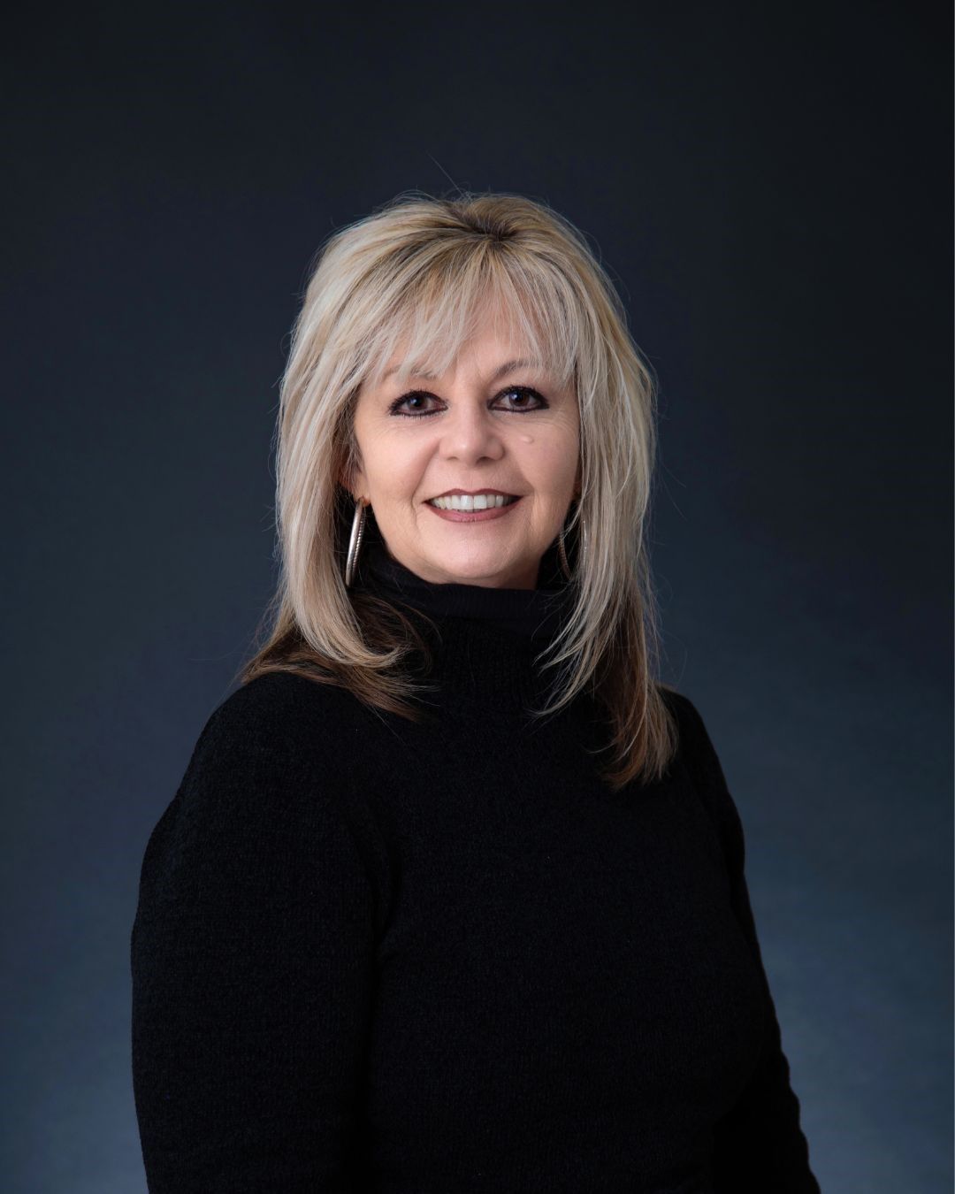 Woman with blonde hair, smiling in a black turtleneck sweater, posing against a dark blue background.